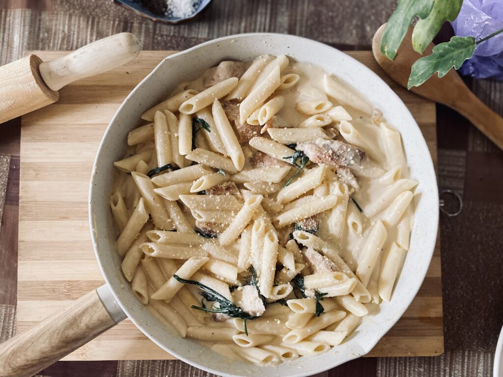 Creamy Chicken Penne: A top view of penne pasta in a creamy sauce, mixed with chicken pieces and garnished with fresh herbs and grated cheese, served in a white skillet on a wooden cutting board.