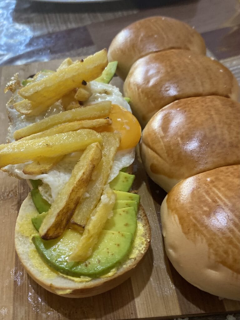 Two caterpillar-shaped breads, one cut in half, is topped with slices of avocado, a sunny-side-up egg, and French fries.