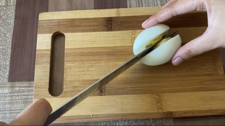 One hand holding an egg in place, the other hand is holding a knife slicing the egg.